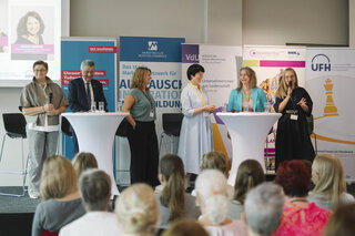 Das Bild zeigt eine Diskussionsrunde mit mehreren Frauen auf einer Bühne, die vor einem Publikum sprechen. Hinter ihnen sind Sponsorenbanner zu sehen.
