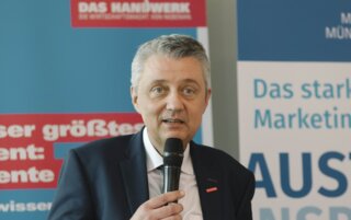 Der Mann spricht mit einem Mikrofon, im Hintergrund Sicht auf Banner von Handwerk und Marketing. Er wirkt engagiert und professionell.