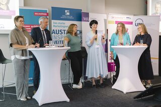 Das Bild zeigt eine Podiumsdiskussion mit mehreren Fachleuten. Sie stehen an Stehtischen, während eine Moderatorin spricht.