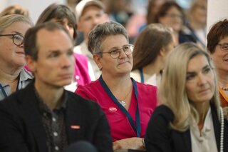 Das Bild zeigt eine Menschenmenge bei einer Veranstaltung. Einige Zuhörer hören aufmerksam zu, während andere beschäftigt wirken.