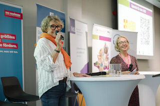 Zwei Frauen stehen an einem Tisch und halten Mikrofone. Sie präsentieren vor einem Publikum, umgeben von Bannern und Plakaten.