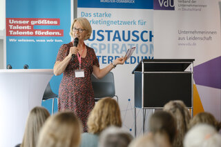 Eine Referentin spricht enthusiastisch vor einem Publikum, umgeben von Bannern, die Talente und Netzwerke fördern.