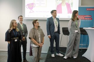 Das Bild zeigt fünf Personen auf einer Veranstaltung, die an einem Podium stehen. Sie wirken aufmerksam und partizipieren aktiv.