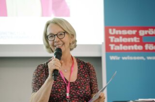 Eine Frau mit Brille und blumenkleid hält ein Mikrofon und spricht auf einer Veranstaltung. Im Hintergrund sind Präsentationsmaterialien sichtbar.