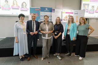 Das Bild zeigt eine Gruppe von sechs Personen, die auf einer Veranstaltung stehen. Im Hintergrund sind Präsentationsfolien zu sehen.