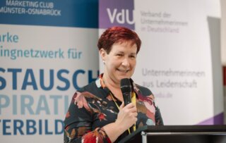 Die Frau am Mikrofon präsentiert in einem Veranstaltungsraum, umgeben von Bannern zweier Organisationen. Sie wirkt engagiert und freundlich.