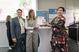 Auf dem Bild stehen drei Personen an einem Stehtisch, während eine von ihnen eine Auszeichnung überreicht. Im Hintergrund ist eine Präsentation zu sehen.