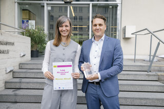 Auf dem Bild stehen zwei Personen vor einem Gebäude. Sie halten Auszeichnungen in der Hand und lächeln in die Kamera.