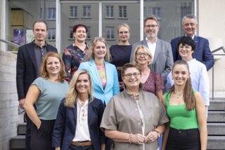 Auf der Treppe stehen mehrere Personen in formeller Kleidung. Sie lächeln in die Kamera, umgeben von einem urbanen Hintergrund.