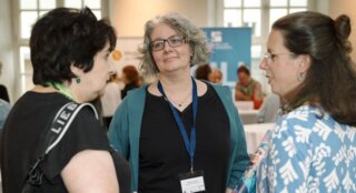 Drei Frauen stehen in einem Gespräch beim Networking-Event. Sie lächeln und wirken interessiert, während im Hintergrund andere Teilnehmende aktiv sind.