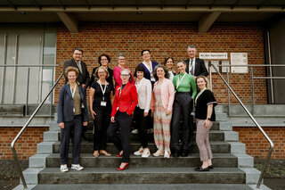 Eine Gruppe von 16 Personen steht auf Treppen vor einem Gebäude. Sie lächeln und tragen verschiedene professionelle Outfits.