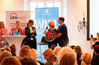 Das Bild zeigt eine Veranstaltung mit Frauen in einem Raum. Eine Person überreicht einem anderen einen Blumenstrauß, während das Publikum applaudiert.