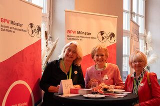 Das Bild zeigt drei Frauen, die an einem Stehtisch stehen und lächeln. Im Hintergrund sind Banner des BPW Münster sichtbar.