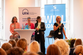 Auf dem Bild stehen mehrere Frauen an einem Podium und sprechen zu einem Publikum. Im Hintergrund sind Banner der Veranstalter sichtbar.