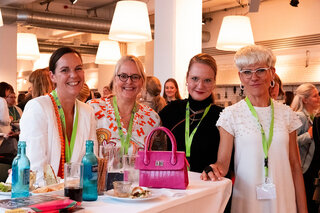 Auf dem Bild sind vier Frauen mit freundlichen Gesichtsausdrücken zu sehen, die an einem Tisch stehen. Sie tragen modische Kleidung und lächeln.