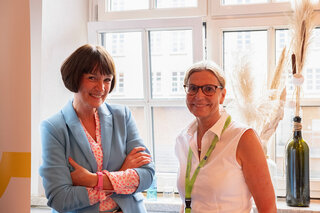 Zwei Frauen stehen lächelnd neben einem Fenster. Eine trägt einen blauen Blazer, die andere eine weiße Bluse. Der Raum hat eine helle, freundliche Atmosphäre.