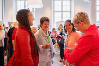 Das Bild zeigt eine Gruppe von Frauen bei einer Veranstaltung. Sie stehen in einem hellen Raum, unterhalten sich und genießen Drinks.