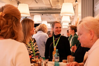 Eine Gruppe von Frauen unterhält sich in einem hellen Raum mit modernen Lampen. Es gibt Getränke und Snacks auf den Tischen. Sie wirken freundlich und interessiert.
