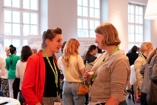 Zwei Frauen unterhalten sich angeregt auf einer Veranstaltung. Im Hintergrund sind weitere Personen und Stände sichtbar. Die Atmosphäre ist lebhaft.