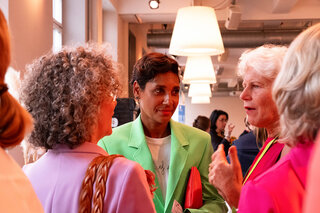 Die Szene zeigt mehrere Frauen in lebhaften Outfits, die in einem modernen Raum miteinander sprechen und lachen. Eine von ihnen trägt ein auffälliges grünes Blazer.