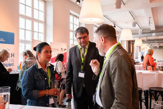 Auf dem Bild stehen drei Personen in einem modernen Raum, offenbar in Gespräche vertieft. Sie tragen Veranstaltungsbadges und wirken interessiert.