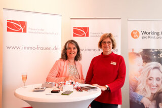Auf dem Bild stehen zwei Frauen an einem Stehtisch, umgeben von Bannern der Initiative „Frauen in der Immobilienwirtschaft“. Ein Glas Sekt steht bereit.