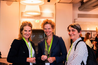 Drei Frauen stehen lächelnd zusammen, halten Gläser in der Hand und tragen farbige Lanyards. Im Hintergrund ist eine moderne Einrichtung zu sehen.
