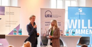 Auf dem Bild sind zwei Frauen auf einer Bühne während einer Veranstaltung zu sehen. Im Hintergrund stehen Banner von verschiedenen Organisationen.