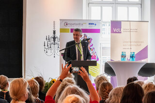 Eine Person steht an einem Rednerpult und spricht vor einem Publikum. Im Hintergrund sind Banner und eine stilvolle Einrichtung zu sehen.