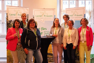 Das Bild zeigt eine Gruppe von Frauen, die an einem Stand mit dem Logo „LandFrauen“ stehen. Sie lächeln und tragen grüne Ausweise.