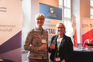 Zwei Frauen stehen in einer Veranstaltungslocation, halten Sektgläser und lächeln. Im Hintergrund sind Banner von verschiedenen Organisationen sichtbar.