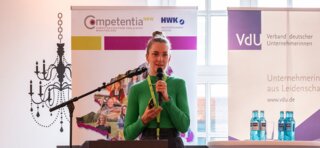 Eine Vortragende mit grüner Bluse steht am Mikrofon und spricht vor einem Publikum, während verschiedene Logos im Hintergrund zu sehen sind.