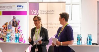 Auf dem Bild sind zwei Frauen auf einer Bühne zu sehen, die vor einem Publikum sprechen. Im Hintergrund stehen Banner von Organisationen und Sponsoren.