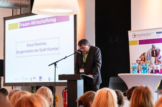 Der Bürgermeister von Münster, Klaus Rosenau, spricht beim Frauen-Wirtschaftstag. Im Hintergrund sind Präsentationsmaterialien sichtbar.