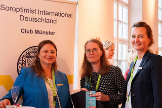 Drei Frauen stehen an einem Soroptimist International Deutschland Banner. Sie lächeln und halten Materialien in den Händen, umgeben von festlicher Atmosphäre.