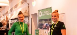 Zwei freundliche Frauen stehen an einem Tisch, lächeln und präsentieren Informationen über "Strong Partners" in einem modernen Eventraum.