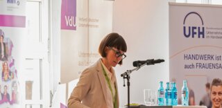 Eine Frau steht am Rednerpult und spricht. Im Hintergrund sind Banner und Stühle zu sehen. Sonnige Sonnenblumen schmücken den Tisch.