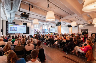 Das Bild zeigt eine große Menschenmenge in einem modernen Raum, die gespannt einem Vortrag folgt, während auf einer Leinwand Informationen angezeigt werden.