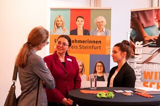 Das Bild zeigt drei Frauen in einem Gespräch vor Bannern, die verschiedene Berufsfelder und Partner anzeigen. Es herrscht eine freundliche Atmosphäre.