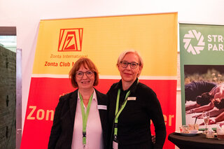 Zwei Frauen stehen lächelnd vor einem Zonta International Banner. Sie tragen Namensschilder und stehen in einem Veranstaltungsraum.