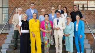 Auf der Treppe stehen 15 Personen in verschiedenen bunter Kleidung. Sie wirken freundlich und lächeln, um eine positive Atmosphäre zu erzeugen.