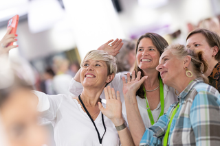 Fünf Frauen machen ein Selfie auf einer Veranstaltung. Sie lächeln, winken und wirken fröhlich und entspannt.