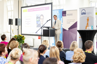 Auf dem Bild ist eine Veranstaltung zu sehen, bei der ein Redner auf der Bühne spricht. Im Publikum sind hauptsächlich Frauen, die interessiert zuhören.