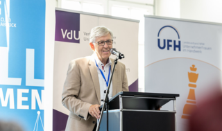 Ein älterer Mann spricht am Mikrofon auf einer Veranstaltung. Im Hintergrund sind Banner von verschiedenen Organisationen zu sehen.