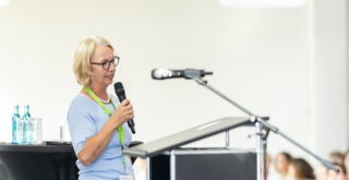 Eine Rednerin steht an einem Pult und spricht zu einem Publikum. Im Hintergrund sind Zuhörer in entspannter Atmosphäre zu sehen.