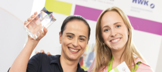 Zwei Frauen stehen lächelnd zusammen, eine hält einen Pokal und die andere ein Zertifikat. Beide strahlen Freude und Stolz aus.