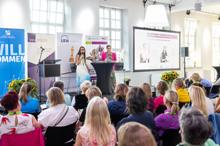 Auf dem Bild sind zwei Frauen auf einer Bühne während einer Veranstaltung zu sehen. Der Raum ist mit Blumen dekoriert und das Publikum hört aufmerksam zu.