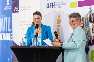 Zwei Frauen stehen an einem Tisch, lächeln und sprechen. Im Hintergrund sind Logos und Banner von Veranstaltungen zu sehen.