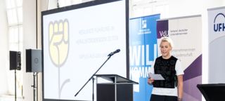 Auf dem Bild spricht eine Frau mit Mikrofon vor einer Präsentation. Im Hintergrund stehen Banner und eine Zuschauergruppe ist sichtbar.