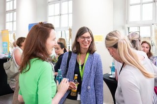 Drei Frauen unterhalten sich angeregt auf einer Veranstaltung, umgeben von fröhlicher Atmosphäre und Getränken. Freundliche Gesichter und lebhafte Gespräche.
